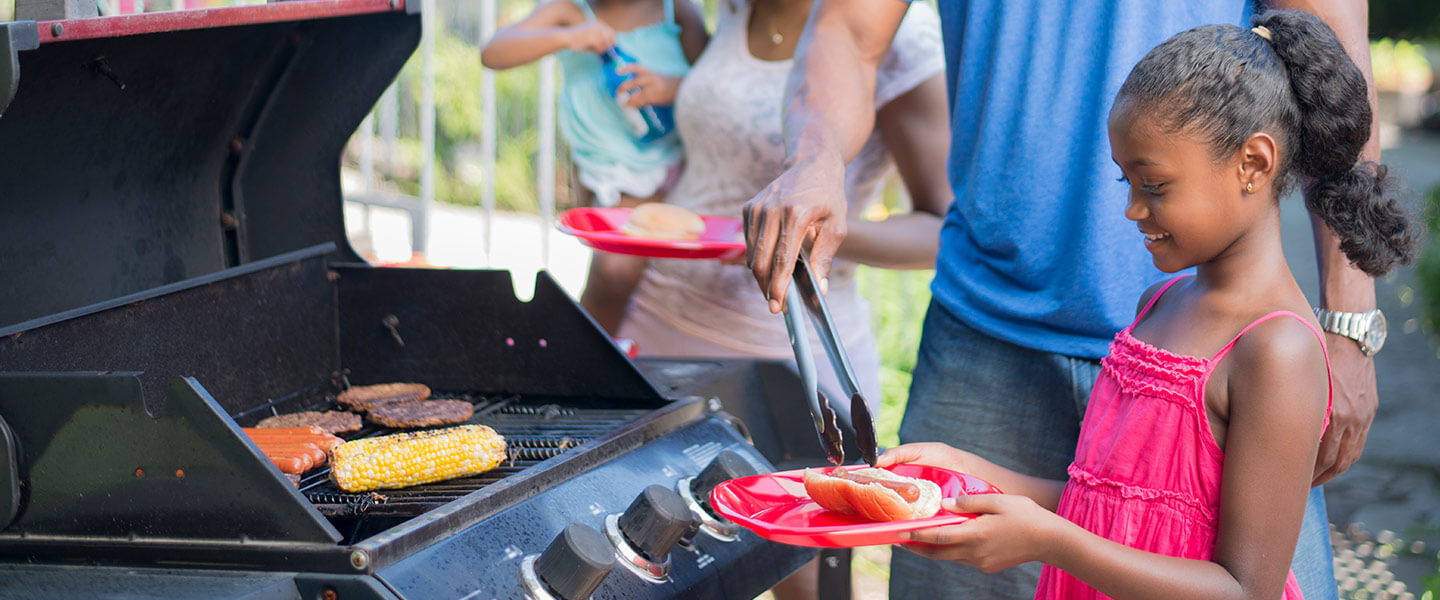 A family of four are outside on a beautiful sunny summer day. They father is barbecuing hot dogs and hamburgers over the grill.