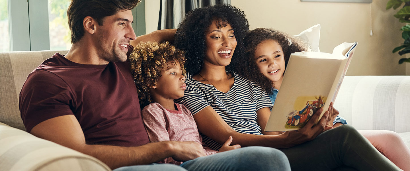 family of four reading a story book together while relaxing on a sofa at home