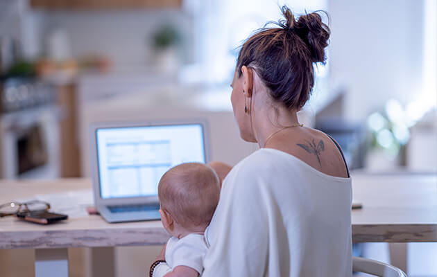 A young Mother sits at the kitchen table with her infant son on her lap as she tries to budget her monthly expenses on her laptop.